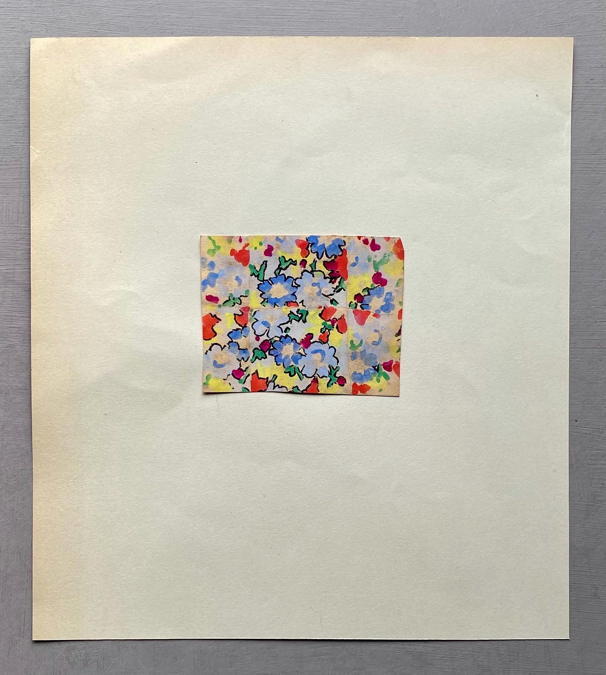 May include: A small, rectangular fabric swatch with a floral pattern of blue, red, and yellow flowers on a cream background. The swatch is centered on a larger, off-white paper sheet. The flowers have green stems and leaves.