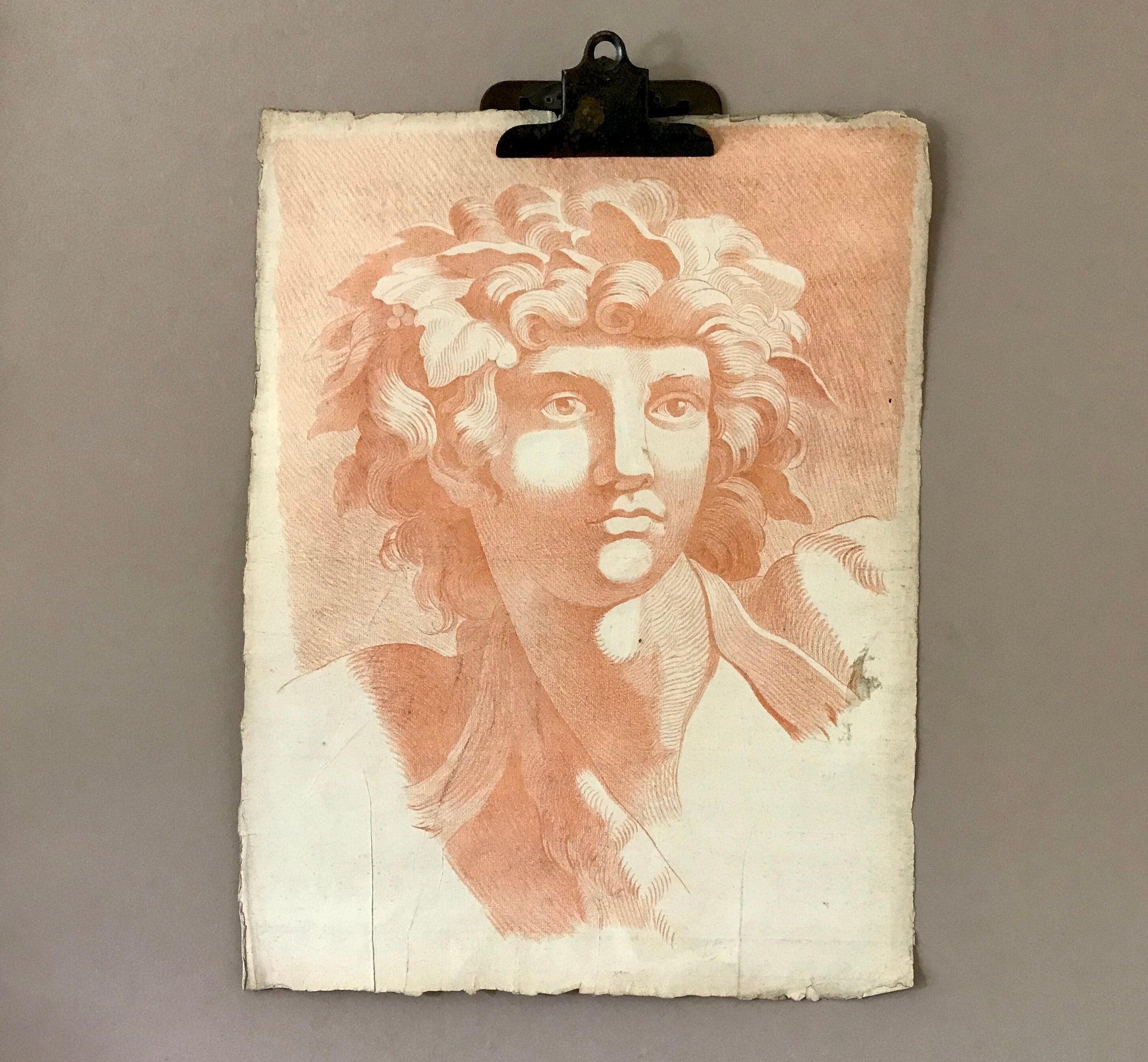 May include: A vintage print of a classical bust, rendered in shades of peach and white. The artwork, held by a metal clip, depicts a figure with curly hair and a serene expression. The paper has a textured, aged appearance, suggesting its antique nature.