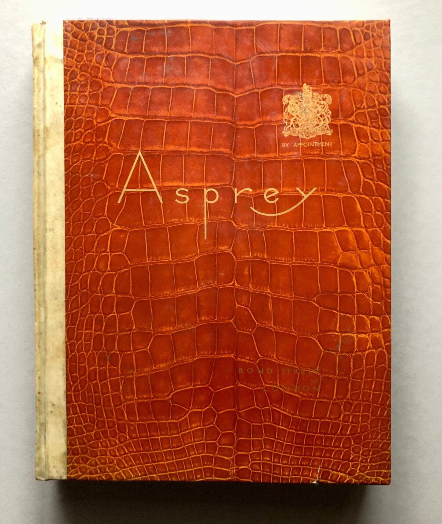 May include: A brown leather-bound book with the word "Asprey" embossed in gold lettering on the front cover. The book also features a gold crest with the text "By Appointment" underneath.