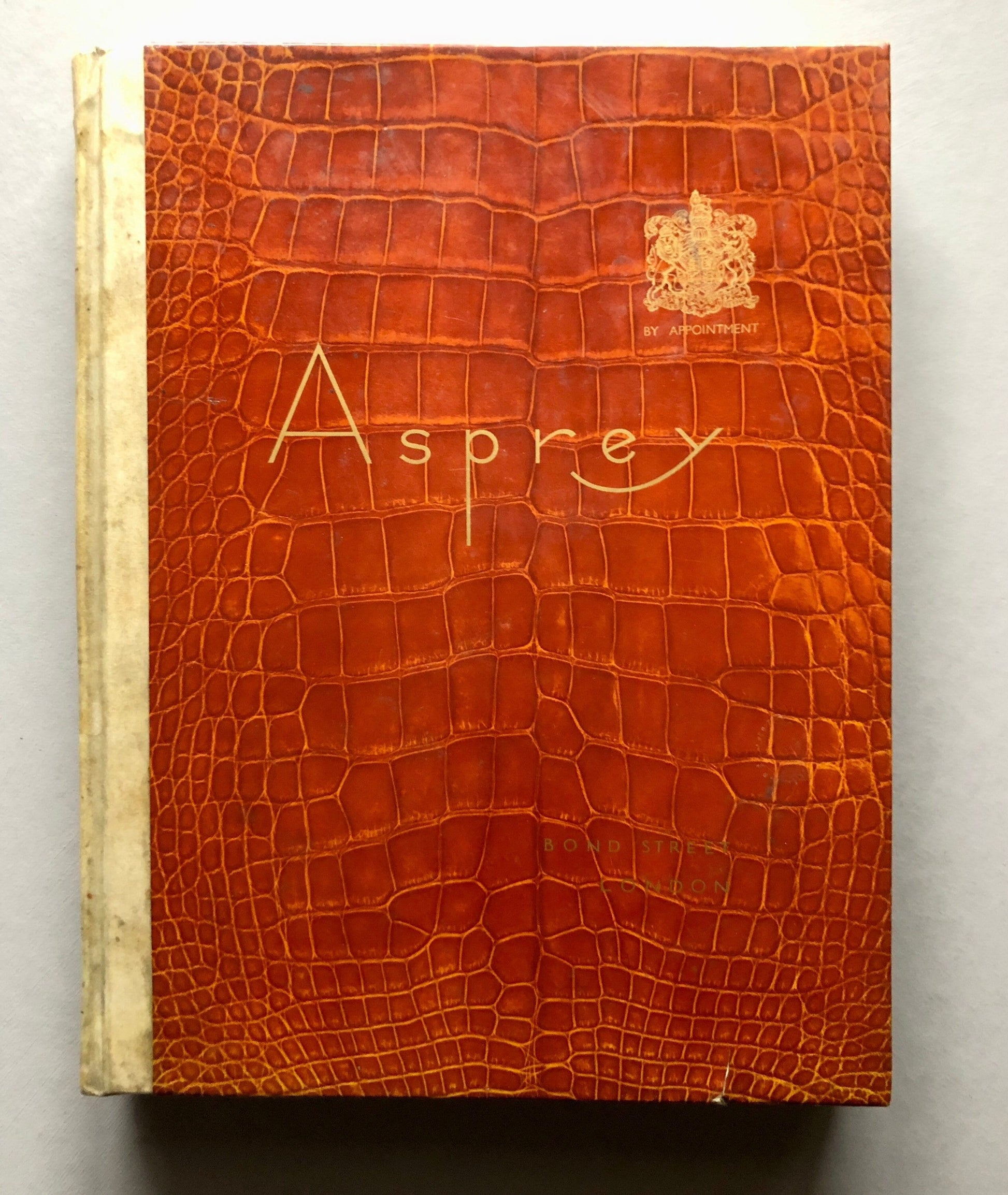 May include: A brown leather-bound book with the word "Asprey" embossed in gold lettering on the front cover. The book also features a gold crest with the text "By Appointment" underneath.